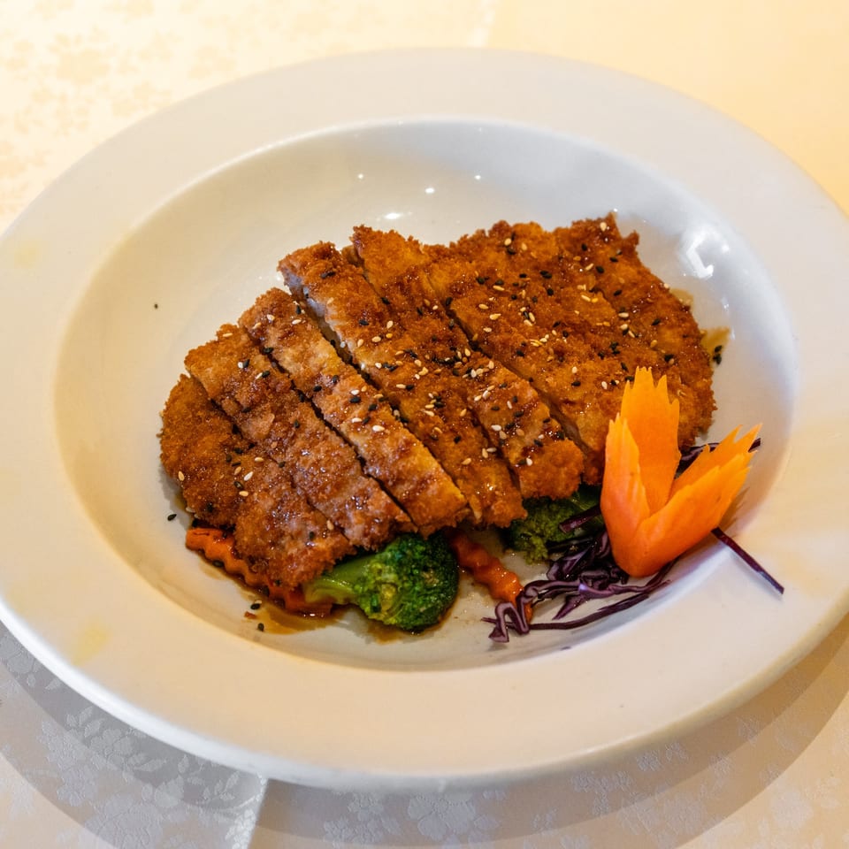 Crispy sliced chicken katsu with sesame seeds, broccoli, and a carved carrot on a white plate.